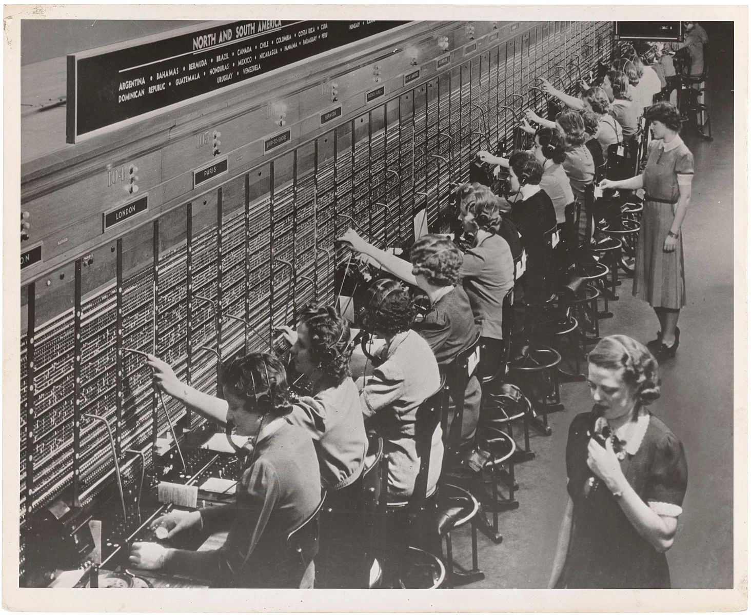A black and white photograph showing a row of women sitting in front of big switchboards